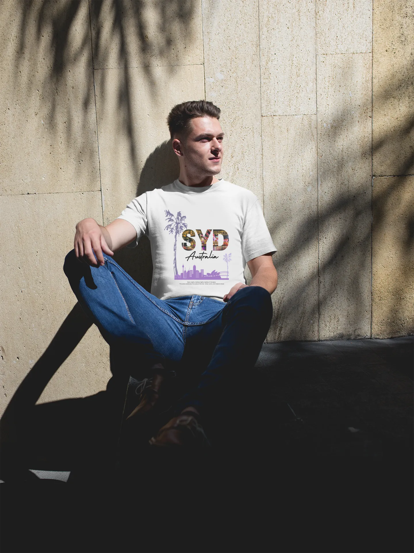 Person wearing a t-shirt with 'SYD' and jeans sitting against a concrete wall.