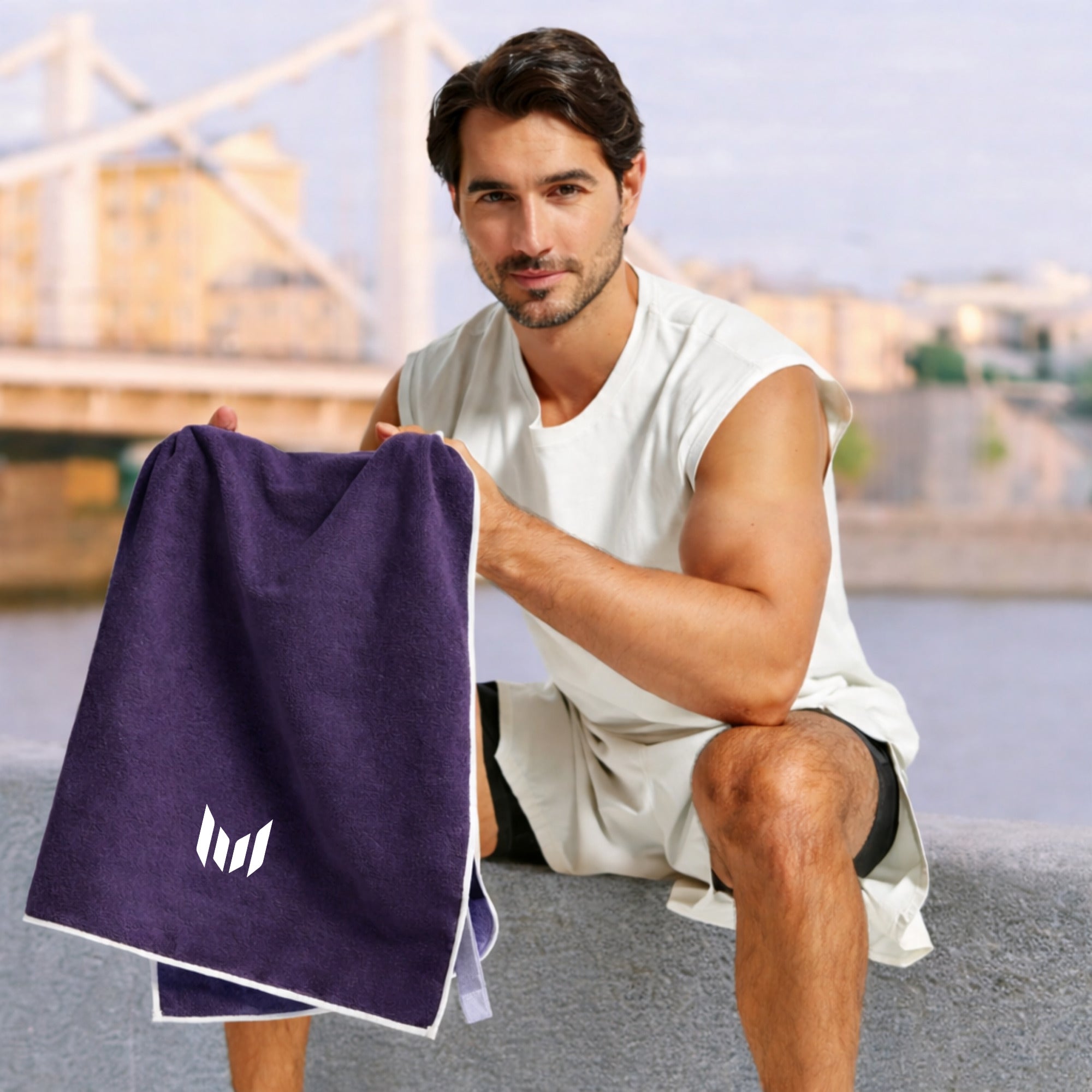 Man holding a purple towel with a logo, sitting outdoors with a blurred cityscape in the background