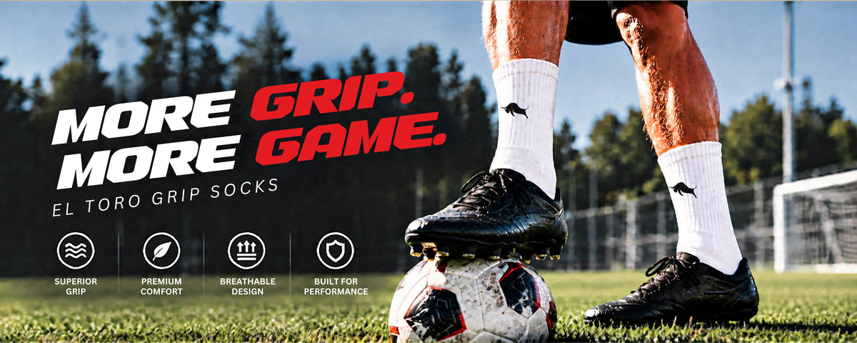 A close-up lifestyle shot of a soccer player's legs and feet on a grass field, wearing white El Toro Grip Socks and black soccer cleats, with a soccer ball on their foot. The background shows a blurred forest and stadium setting.