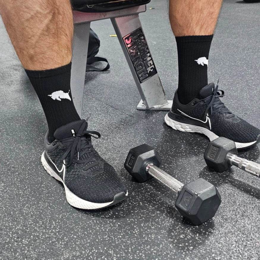 Person wearing black athletic shoes and El Toro black grip socks with a logo, standing next to dumbbells on a gym floor.