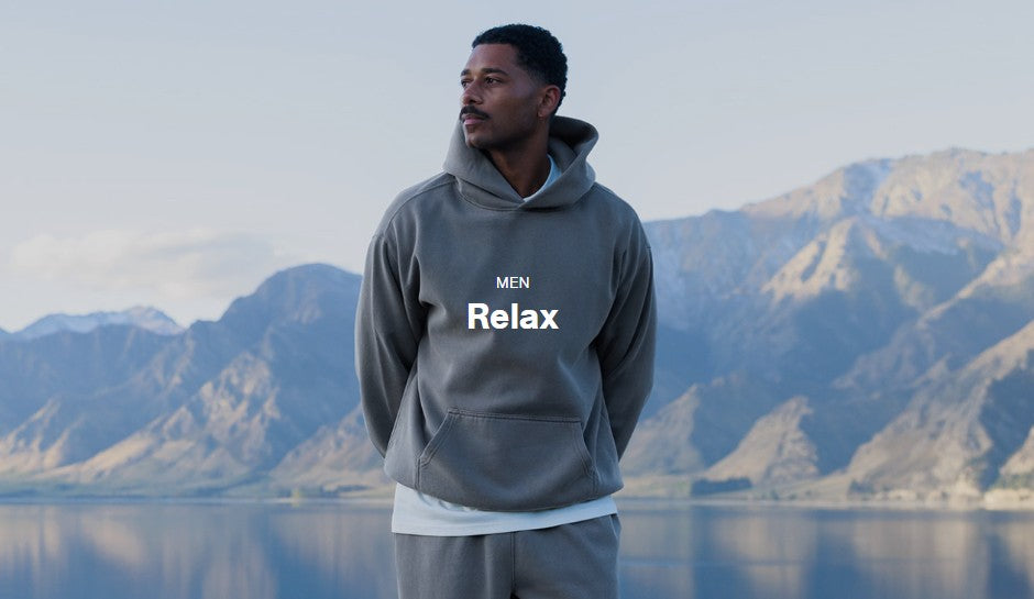 A man wearing a gray pullover hoodie with 'MEN Relax' printed on the chest stands outdoors in front of a mountain and lake landscape.
