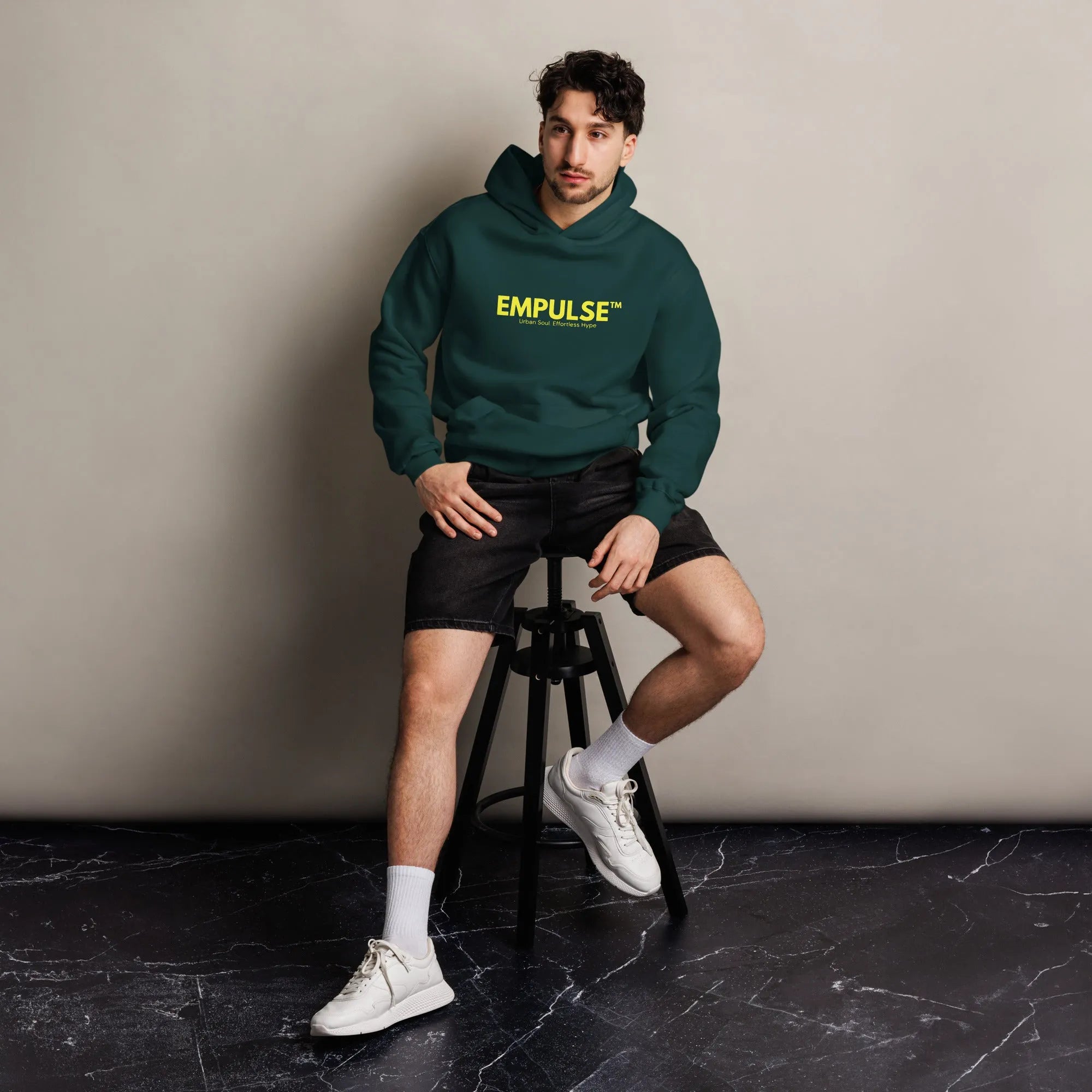 Young man seated in Empulse™ green hoodie with yellow branding, minimal industrial backdrop