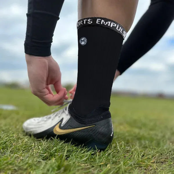 Athlete lacing boots while wearing black compression socks on grass field with soccer boots