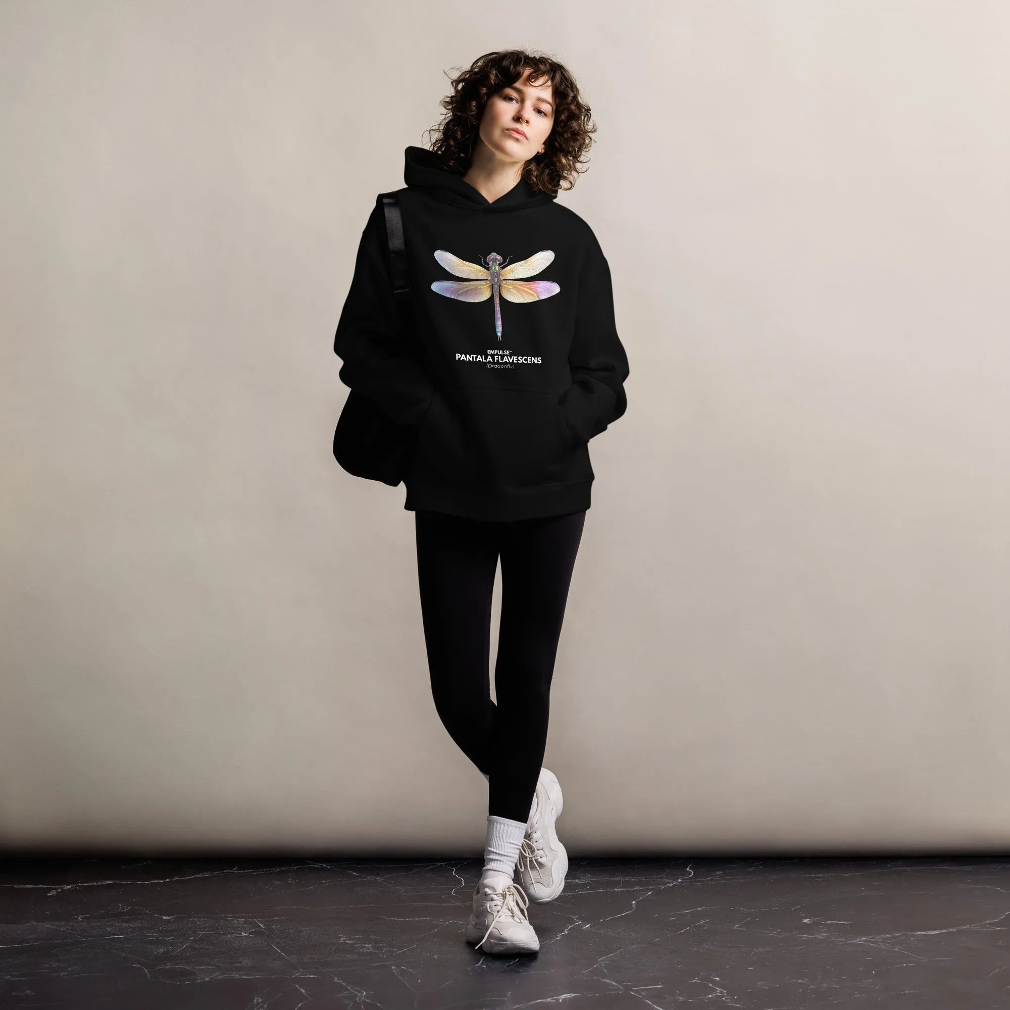 Woman in Empulse™ Dragonfly hoodie leaning against a studio wall in minimalist setting

